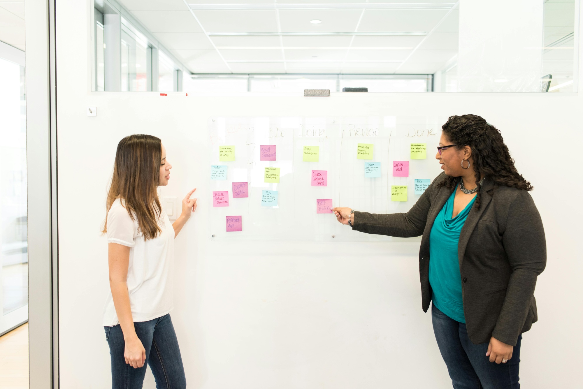 Two ladies standing in front of a whiteboard discussing post-it notes that are stuck on it